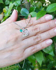 Hand displaying a round cut emerald dolphin pendant against a green leafy background.