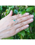 Hand displaying a round cut emerald dolphin pendant against a green leafy background.