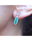 14K white gold dangling earrings with rectangular emeralds and diamond clusters on an ear.