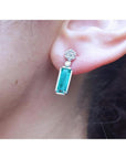 14K white gold dangling earrings with rectangular emeralds and diamond clusters on an ear.