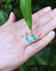 Hand displaying a pair of emerald earrings with a leafy background