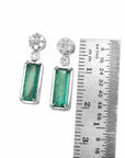 Pair of emerald earrings with accent cluster diamonds next to a ruler for size reference on a white background.