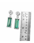Pair of emerald earrings with accent cluster diamonds next to a ruler for size reference on a white background.