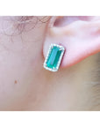 Emerald-cut Colombian emerald stud earrings with a diamond halo, set in 18K gold on an earlobe.
