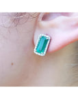 Emerald-cut Colombian emerald stud earrings with a diamond halo, set in 18K gold on an earlobe.