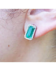 Emerald-cut Colombian emerald stud earrings with a diamond halo, set in 18K gold on an earlobe.