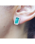 Emerald-cut Colombian emerald stud earrings with diamond halo in 18K gold, worn on an ear.