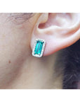 Emerald-cut Colombian emerald stud earrings with diamond halo in 18K gold, worn on an ear.