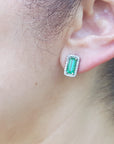 Close-up of an ear wearing a green emerald-cut earring with a halo diamond and white gold setting.
