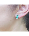 Close-up of an ear wearing a green emerald-cut earring with a halo diamond and white gold setting.