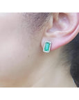 Emerald-cut emerald stud earrings with diamond halo, worn on an ear, in 18K gold.