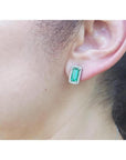 Emerald-cut emerald stud earrings with diamond halo, worn on an ear, in 18K gold.