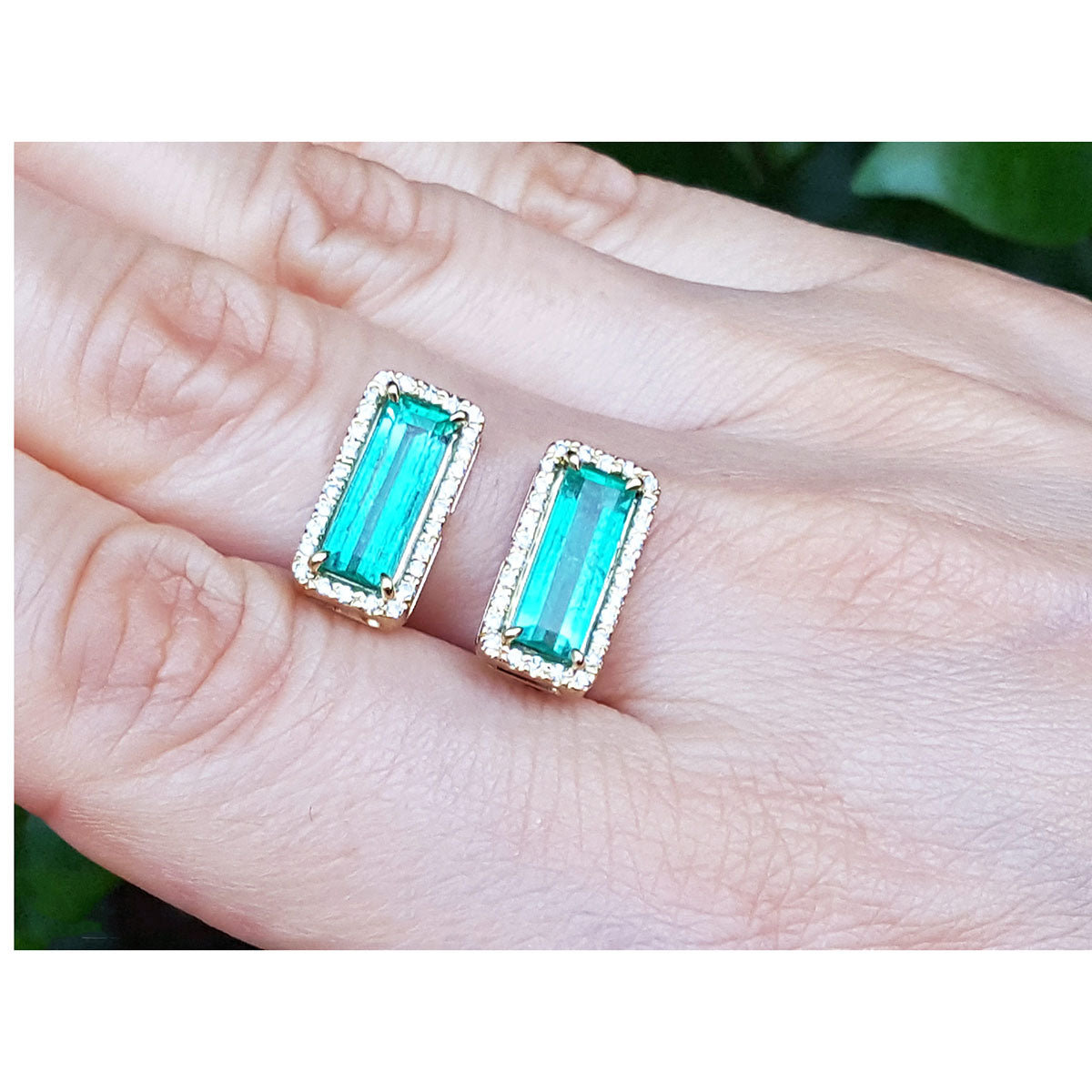 Emerald gemstone earrings held between fingers against a neutral background