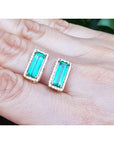 Emerald gemstone earrings held between fingers against a neutral background