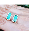 Emerald gemstone earrings held between fingers against a neutral background