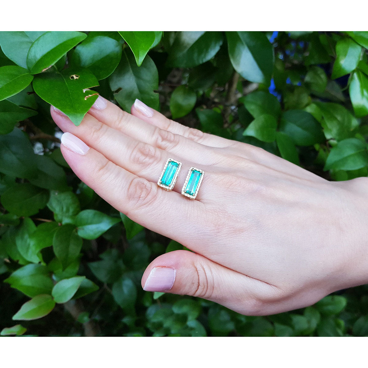 Hand holding two emerald green earrings against a green leafy background