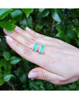 Hand holding two emerald green earrings against a green leafy background