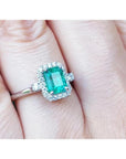 An inexpensive emerald engagement ring with a halo diamond white gold on a finger.
