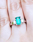 A Colombian emerald engagement ring on a finger against a natural background.