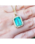 Emerald-cut emerald pendant with halo diamonds in 18K gold, worn on a hand.