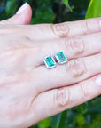 A pair of emerald and diamond Colombian emerald stud earrings on a hand with a blurred green background.