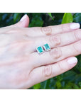 Emerald-cut natural Colombian emerald stud earrings with diamond halo, set in 14K white gold, on a hand.