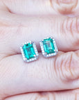 Emerald green emerald-cut gemstone earrings with diamond halo on a hand.
