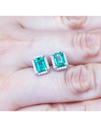 Emerald-cut natural Colombian emerald stud earrings with a halo of diamonds in 14K gold, held on a hand.