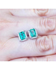 Emerald-cut natural Colombian emerald stud earrings with a halo of diamonds in 14K gold, held on a hand.