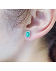 Emerald-cut natural emerald stud earrings with a halo of diamonds set in 14K gold on an ear.