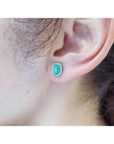 Emerald-cut natural emerald stud earrings with a halo of diamonds set in 14K gold on an ear.