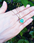 Hand holding a gold necklace with a natural emerald heart pendant against a leafy background.