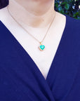 A woman wearing a blue top with a necklace featuring a natural colombian emerald heart pendant.