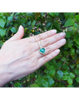 Emerald heart pendant with diamond halo on an 18k gold chain, held against green foliage.