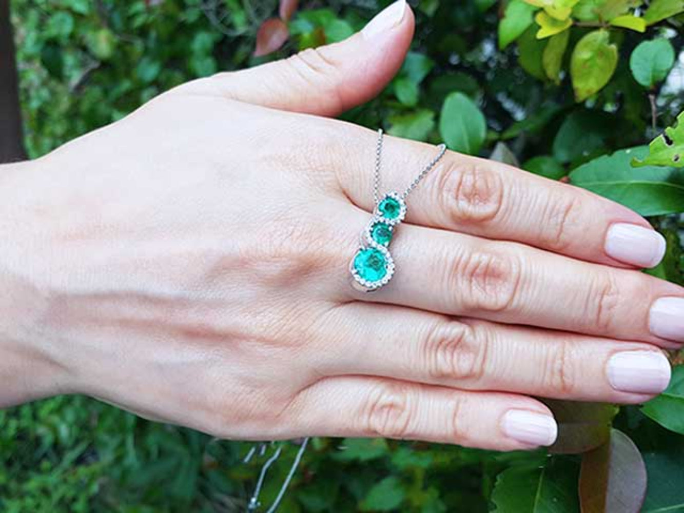 Hand displaying a white gold journey emerald pendant necklace against a green leafy background.