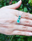 Hand displaying a white gold journey emerald pendant necklace against a green leafy background.