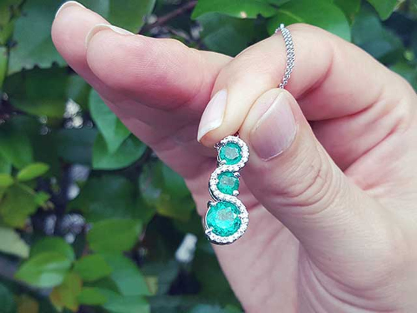 Hand holding between fingers an emerald journey pendant necklace with a blurred green leafy background.