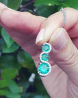 Hand holding between fingers an emerald journey pendant necklace with a blurred green leafy background.