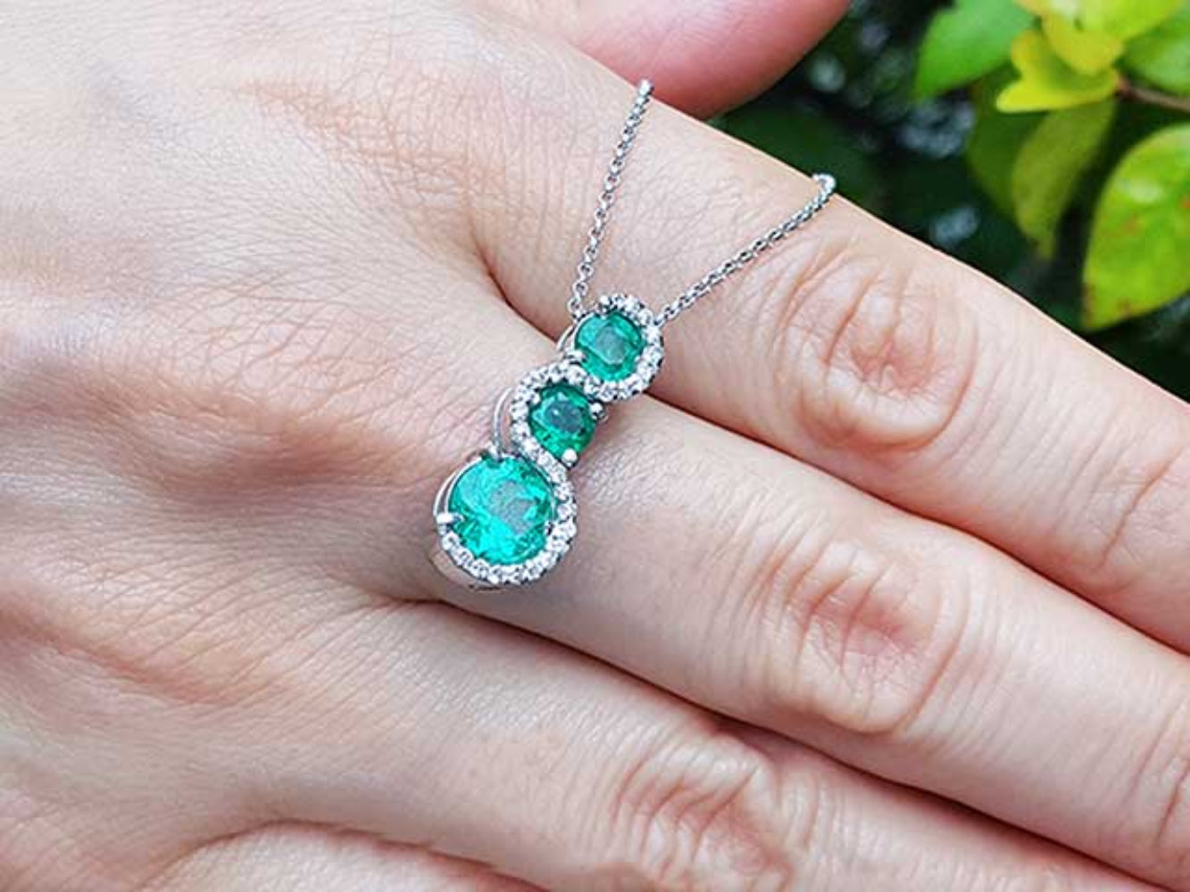 Emerald journey pendant necklace on a hand with a blurred green background.