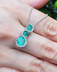 Emerald journey pendant necklace on a hand with a blurred green background.