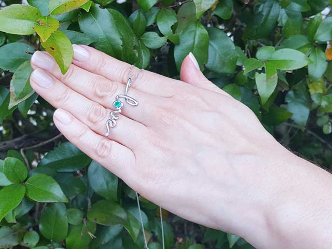 Hand displaying a 14K solid gold emerald love pendant against a green leafy background.