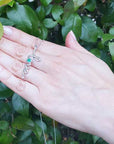 Hand displaying a 14K solid gold emerald love pendant against a green leafy background.