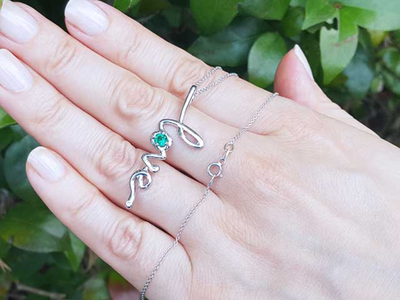 A 14K solid gold emerald love pendant on a hand against a green leafy background.