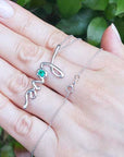 A 14K solid gold emerald love pendant on a hand against a green leafy background.