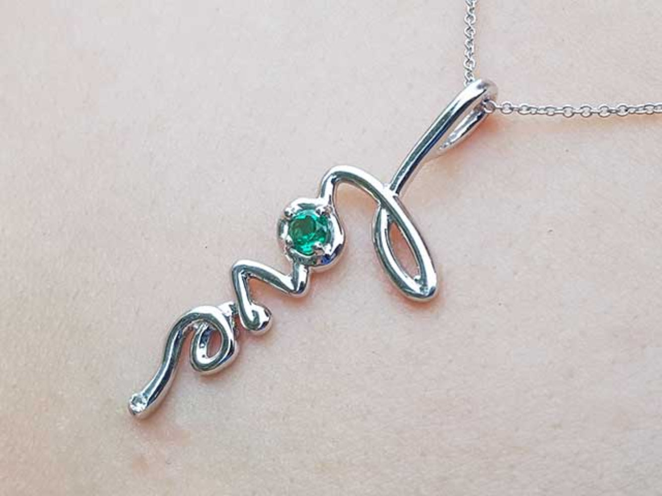 A white gold script emerald love pendant on a light pink skin background.