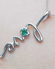 A white gold script emerald love pendant on a light pink skin background.