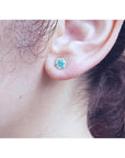 14K white gold stud earring with a round teal gemstone surrounded by tiny white stones, worn on an ear.