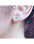 14K white gold stud earring with a round teal gemstone surrounded by tiny white stones, worn on an ear.