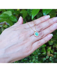 Hand displaying a yellow gold necklace with a oval cut emerald pendant against a green leafy background