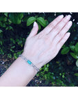 14K white gold paperclip bracelet with a vibrant emerald-cut Colombian emerald and diamond halo on a woman's wrist.